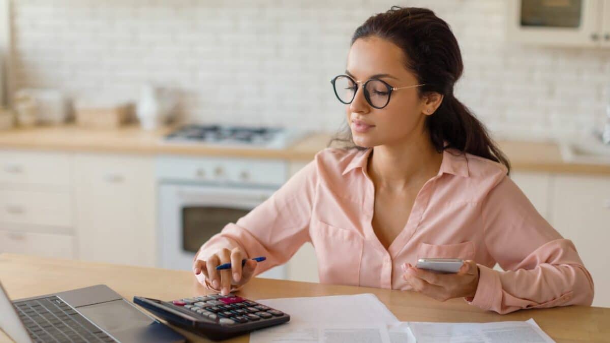 Jeune femme concentrée portant des lunettes utilisant un smartphone et une calculatrice devant une table de cuisine en bois, entourée de documents