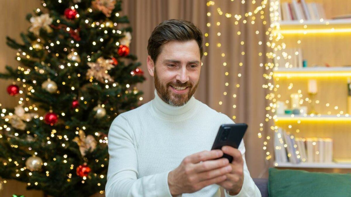 Un homme joyeux à la maison utilise le téléphone, célèbre Noël et le Nouvel An assis sur le canapé près du sapin